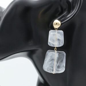 May include: Gold-tone earrings with a post and a large, round stud. The earrings feature two square, marbled, white and gray acrylic pendants suspended by a thin gold-tone wire. The earrings are displayed on a black mannequin head.