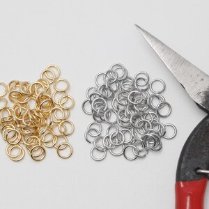 Op de afbeelding: Close-up van benodigdheden voor het maken van sieraden. Twee stapels metalen sprongringen, één goudkleurig en één zilverkleurig, liggen naast een tang met rode handvatten. De sprongringen worden gebruikt voor het maken van sieraden en andere ambachten.