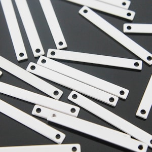 Op de afbeelding: Een groep zilveren metalen staafhangers met twee gaten op elk. De hangers zijn verspreid over een zwart oppervlak.
