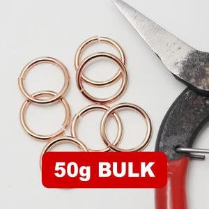 Op de afbeelding: Een verzameling roségouden sprongringen en een metalen schaar met rode handvatten. Een rood rechthoek met de tekst "50g BULK" is in de afbeelding. De sprongringen liggen op een witte ondergrond.