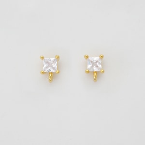 May include: Two gold-colored stud earrings. Each earring features a square, clear cubic zirconia stone set in a gold-colored frame. The earrings have a post and loop design for attachment.