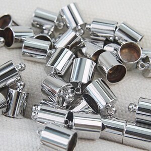 Op de afbeelding: Een stapel zilverkleurige metalen eindkappen voor koorden of sieraden. De doppen hebben een cilindrische vorm met een kleine lus aan de bovenkant.