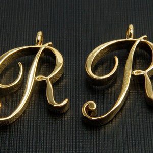 May include: Two gold-colored metal charms in the shape of the letter R. The charms are in a cursive font and have a textured surface.