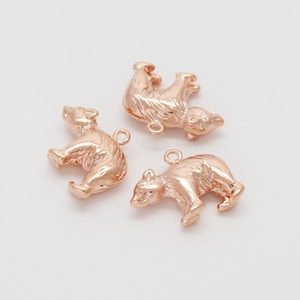Peut inclure: Trois breloques ours en or rose dans différentes poses. Chaque breloque a une petite boucle en haut pour l'attacher à un bijou. Les ours sont détaillés avec une fourrure texturée et sont posés sur un fond blanc.