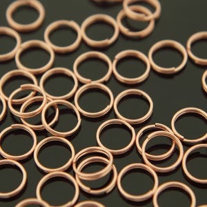May include: A scattering of small, copper-coloured split rings on a dark surface. The rings are circular with a split, allowing them to be opened and attached. Commonly used in jewellery making and crafts.