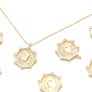 May include: A gold necklace with a pendant featuring a sunburst design with a letter J in the center. The pendant is surrounded by small clear stones. There are six additional pendants with the same design, each with a different letter.