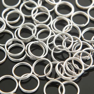 Könnte beinhalten: Ein Haufen silberner Schlüsselringe, auch bekannt als Splintringe, liegen auf einer schwarzen Oberfläche verstreut. Die Ringe sind aus Metall und dienen zum Anbringen von Schlüsseln, Anhängern oder anderen kleinen Gegenständen.