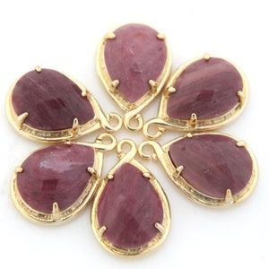 May include: Six teardrop-shaped pendants with a gold-colored frame and a deep red-purple stone. The pendants are arranged in a circular pattern against a white background. Each stone has a faceted surface.