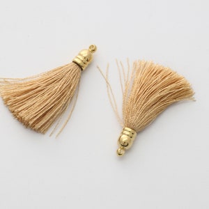 May include: Two beige tassels with gold metal tops. The tassels are made of soft, fine threads and have a delicate, elegant look.