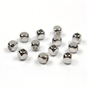 May include: A group of 12 silver metal beads with a square shape and a hole in the center. The beads are scattered on a white surface.