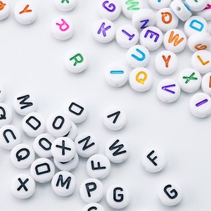 May include: White round beads with black letters and colorful letters scattered on a white surface. The letters are in various colors, including orange, purple, green, and blue. The beads are for crafts.