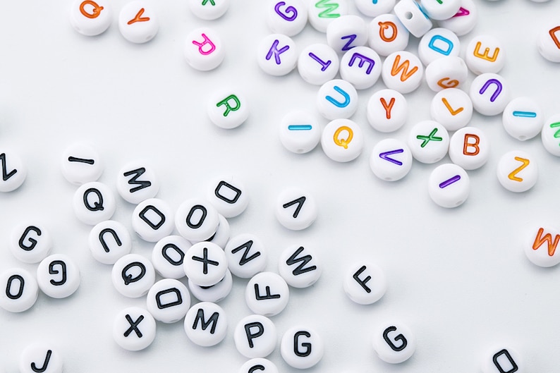 May include: White round beads with black letters and colorful letters scattered on a white surface. The letters are in various colors, including orange, purple, green, and blue. The beads are for crafts.