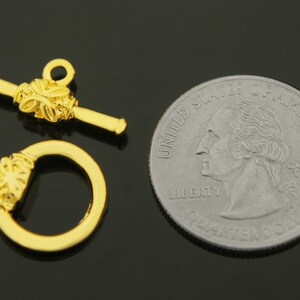 May include: Gold-toned metal toggle clasp with a decorative floral design. The clasp is shown next to a US quarter for size comparison.