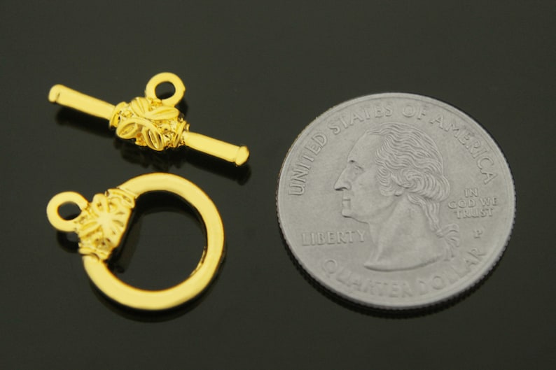 May include: Gold-toned metal toggle clasp with a decorative floral design. The clasp is shown next to a US quarter for size comparison.