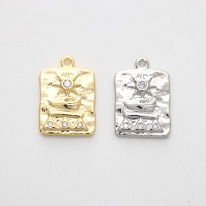 May include: Two gold and silver charm pendants with a textured rectangular design. Each pendant features a raised image of a stylized sun with a star in the center and a small boat below. The pendants are adorned with five clear rhinestones.