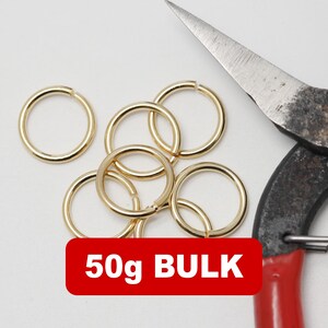 Op de afbeelding: Een verzameling goudkleurige sprongringen met een vintage metalen schaar. Een rode rechthoek met witte tekst leest "50g BULK". De sprongringen zijn verspreid op een wit oppervlak.