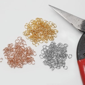 Op de afbeelding: Een bovenaanzicht toont drie stapels metalen verbindingsringen in goud, roségoud en zilver, naast een vintage schaar met rode handvatten. De verbindingsringen worden gebruikt voor het maken van sieraden en andere ambachten.