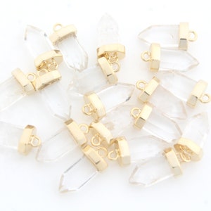 Op de afbeelding: Een groep van heldere kwartskristal hangers met goudkleurige springringen. De hangers zijn gevormd als lange, dunne punten.