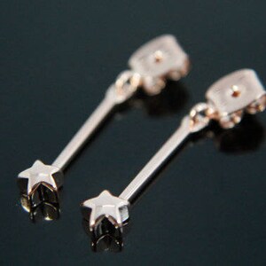 Puede incluir: Un par de pendientes colgantes de estrella de color dorado. Los pendientes tienen un diseño sencillo con un pequeño dije de estrella que cuelga de una varilla delgada.