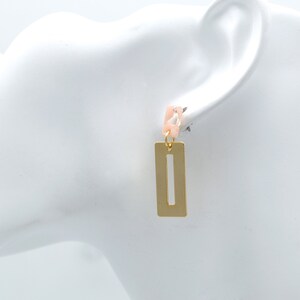 Peut inclure: Boucles d'oreilles rectangulaires dorées avec une découpe verticale. Les boucles d'oreilles ont une petite partie supérieure rose clair et blanche. Les boucles d'oreilles sont présentées sur une tête de mannequin blanche.