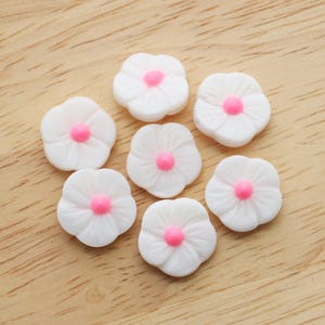 Peut inclure: Sept ornements blancs en forme de fleur avec des centres roses sont disposés sur une surface en bois brun clair. Les fleurs ont un design délicat en forme de pétale, et les centres roses ajoutent une touche de couleur.