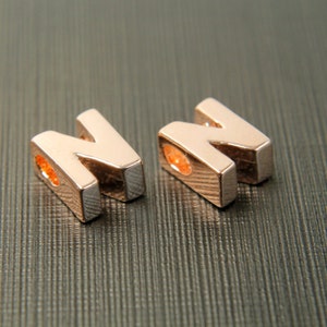May include: Two rose gold letter "N" charms. Each charm has a rectangular shape with a rounded top and a hole for stringing. The charms are displayed on a textured, dark gray surface.