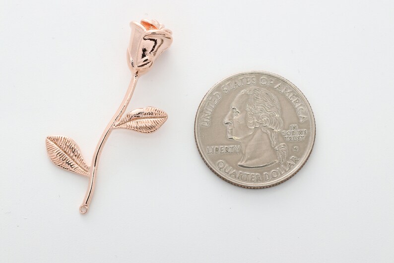 May include: Rose gold rose charm pendant with a single leaf on each side. The charm is shown next to a US quarter for size comparison.