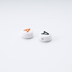 May include: Two white, round beads with a hole drilled through the center. One bead has an orange letter "A", and the other has a black letter "A". The beads are on a white background.