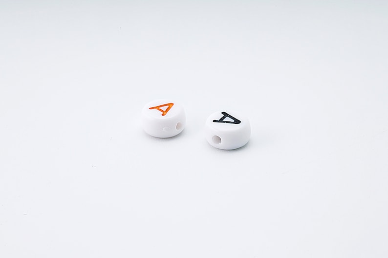 May include: Two white, round beads with a hole drilled through the center. One bead has an orange letter "A", and the other has a black letter "A". The beads are on a white background.
