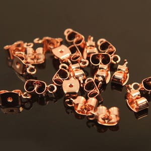 Puede incluir: Primer plano de un grupo de cierres de pendientes de oro rosa. Los cierres son de metal y tienen un pequeño bucle en un lado y un pequeño gancho en el otro. Están diseñados para ser usados con pendientes de botón.