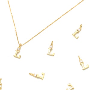 May include: Gold-colored initial "L" pendant necklace with a small, clear gemstone. Several loose "L" charms are scattered around the necklace on a white background. The necklace chain is delicate and twisted.
