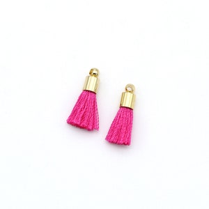 May include: Two small, bright pink tassels with gold-coloured metal caps and loops. The tassels are made of tightly wound threads, and the caps have a smooth, shiny finish. The background is plain white.
