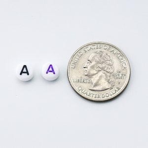 May include: Two white round beads with the letter "A" in black and purple, next to a U.S. quarter coin. The beads are small, likely used for jewelry or crafts. The quarter provides a size reference.