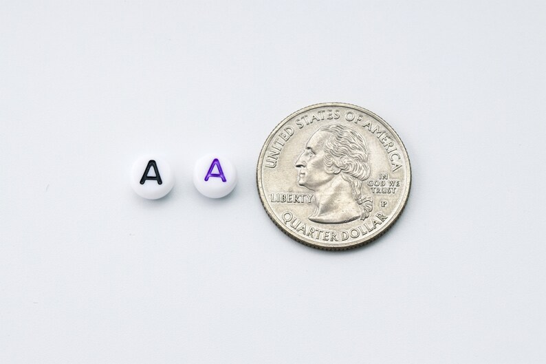 May include: Two white round beads with the letter "A" in black and purple, next to a U.S. quarter coin. The beads are small, likely used for jewelry or crafts. The quarter provides a size reference.