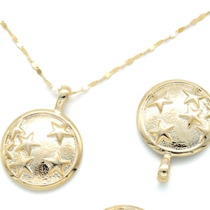 May include: Gold-colored necklace with a pendant featuring a circular design with embossed stars. The necklace has a delicate chain and is set against a white background. Several star-shaped charms are also visible.