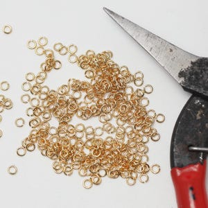 May include: A collection of small, gold-colored jump rings scattered on a white surface, alongside a pair of well-used metal pliers with red handles. The jump rings are used for jewelry making and crafting.