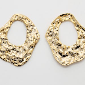 Könnte beinhalten: Zwei goldfarbene Ohrringe in abstrakter Form mit unregelmäßiger, strukturierter Oberfläche. Die Ohrringe haben eine ovale Öffnung in der Mitte und ein glänzendes Finish. Der Schmuck wird auf weißem Hintergrund präsentiert.