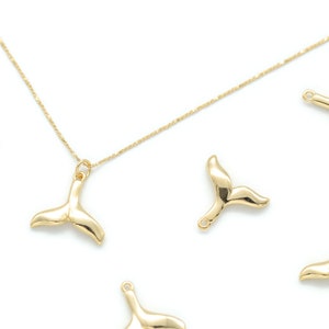 Könnte beinhalten: Goldfarbene Walflossen-Anhänger-Halskette und -Anhänger. Die Halskette hat eine zarte Kette mit einem Walflossen-Anhänger. Mehrere Walflossen-Anhänger sind auf weißem Hintergrund verstreut. Die Anhänger haben eine Schlaufe zum Befestigen an Schmuck.