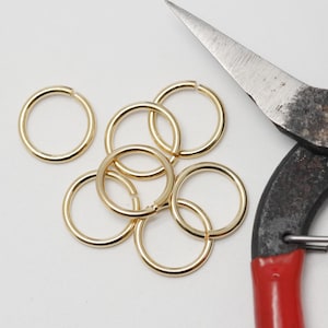 Op de afbeelding: Een verzameling goudkleurige sprongringen en een vintage metalen tang met rode handgrepen. De sprongringen zijn cirkelvormig. De tang is verouderd en vertoont gebruikssporen.