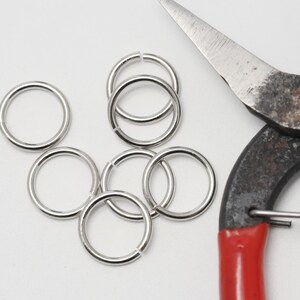 Op de afbeelding: Close-up van zilveren sprongringen en een metalen schaar met rode handvatten. De sprongringen liggen verspreid op een witte ondergrond.