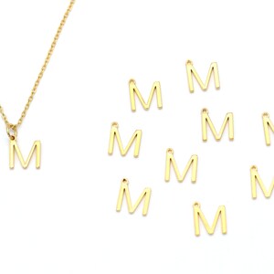 May include: A gold-colored necklace with a pendant in the shape of the letter "M" is displayed. Several individual gold "M" charms are scattered nearby. The jewelry is set against a white background.