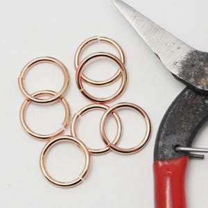 Op de afbeelding: Een verzameling roségouden sprongringen en een vintage tang met rode handgrepen. De sprongringen zijn gerangschikt op een wit oppervlak, geschikt voor het maken van sieraden of knutselprojecten.