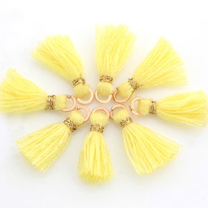 May include: Eight yellow tassels with gold-coloured wire wrapping and copper-coloured jump rings. The tassels are arranged in a circular pattern against a white background. Suitable for jewellery making or craft projects.