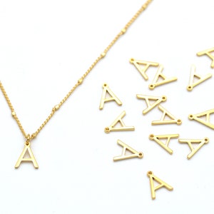 May include: A gold necklace with a small gold letter A charm. There are several additional letter A charms scattered on a white surface.