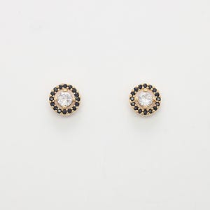 Op de afbeelding: Een paar gouden oorbel studs met een kleine ronde, heldere steen omgeven door een zwarte halo.