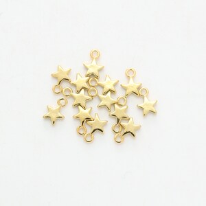 May include: Gold-coloured star charms with small loops for jewellery making. The charms are scattered on a white surface. These star-shaped pendants are suitable for necklaces, bracelets, and earrings.