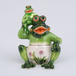 May include: A whimsical green frog figurine with a smaller frog perched on its head. The larger frog has an open mouth and orange accents. The base is white with purple flower details. A decorative item.
