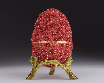 Puede incluir: Un huevo rojo adornado con joyas con un borde dorado que se asienta sobre un soporte de metal dorado con tres patas. El huevo está cubierto de pequeñas rosas rojas.