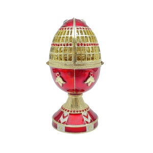 May include: A decorative red and gold egg-shaped ornament with a lattice top and a pedestal base. The egg features gold accents, red jewels, and a gold cage design. The base has a red and gold pattern.