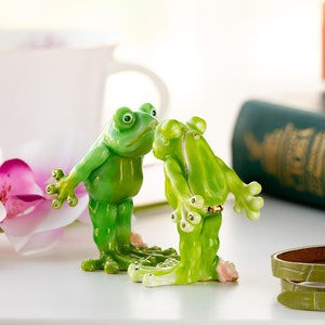 May include: Two green frog figurines facing each other, one with its arms outstretched and the other with its arms crossed, on a white surface.